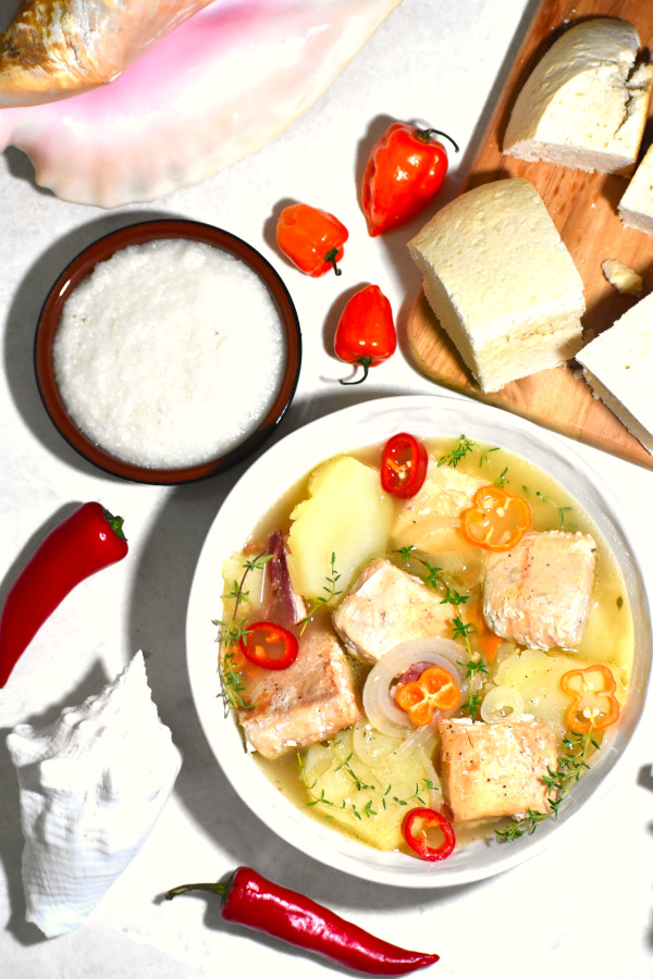 zoomed out image of a bowl of boiled fish alongside some johnny cake, grits and a few tropical shells